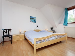Quarto com cama de madeira, duas mesas de cabeceira, uma cadeira, uma pequena mesa, luminária na parede e janela com cortinas.