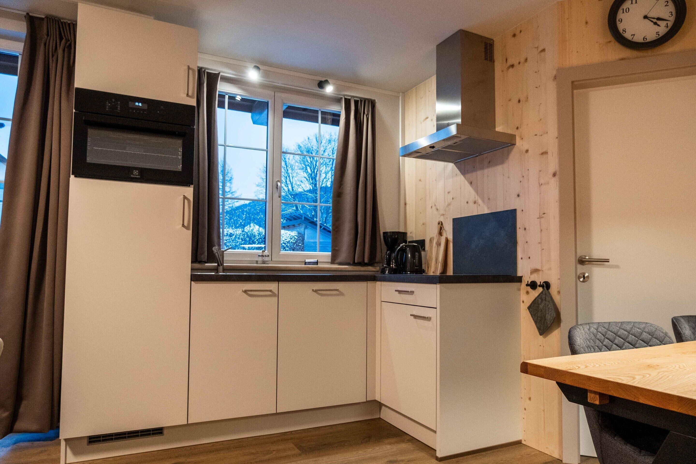 A compact kitchen with a built-in oven, a window with curtains, a wooden wall, and a dining table with chairs.