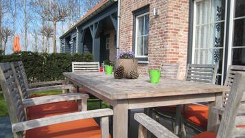 Une table en bois placée à l’extérieur, avec des chaises, un mur en briques et une fenêtre avec des rideaux.