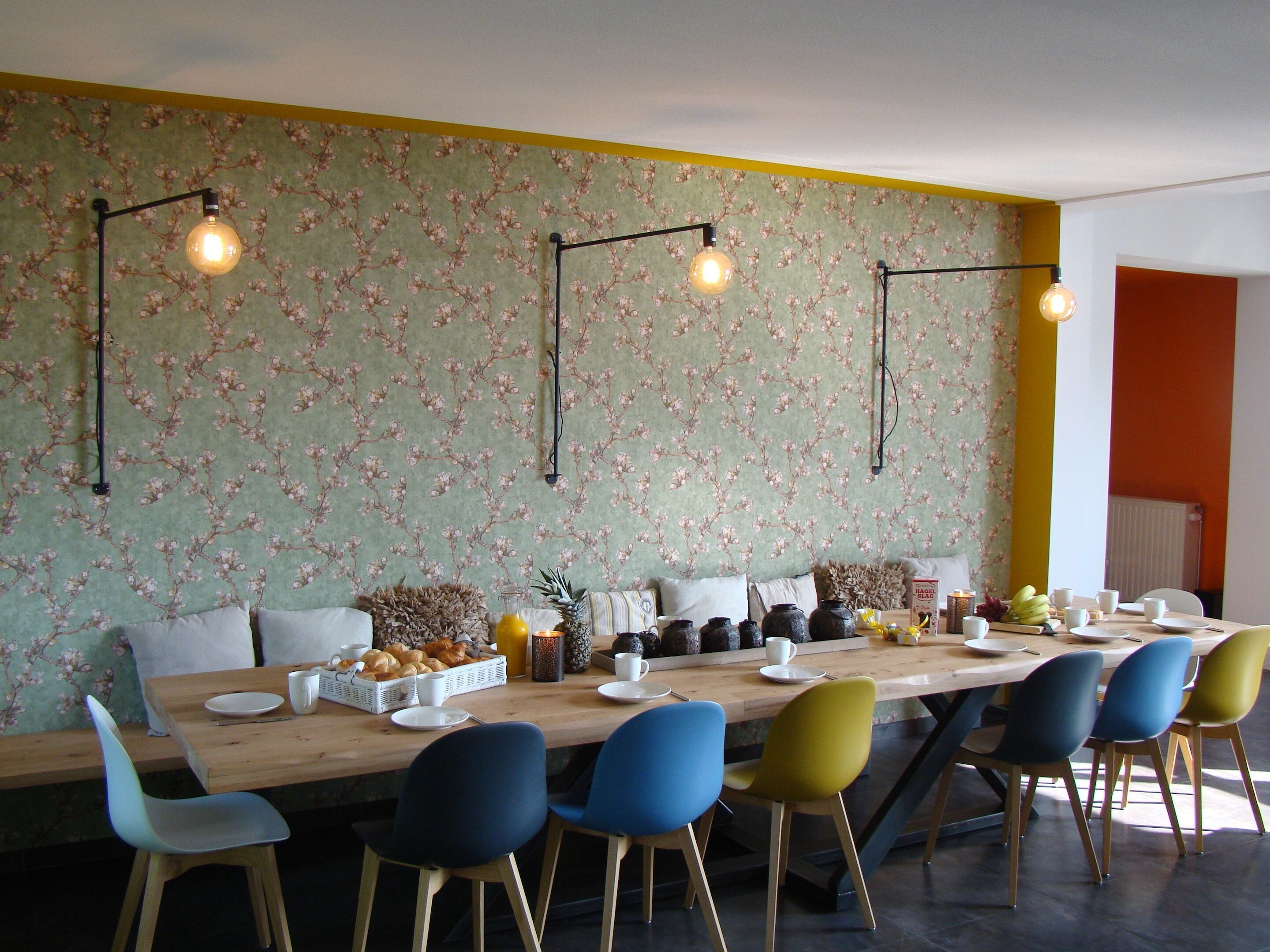 Un espace repas avec une longue table en bois, des chaises colorées, un papier peint à motifs floraux et des luminaires suspendus.