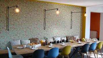 Un espace repas avec une longue table en bois, des chaises colorées, un papier peint à motifs floraux et des luminaires suspendus.