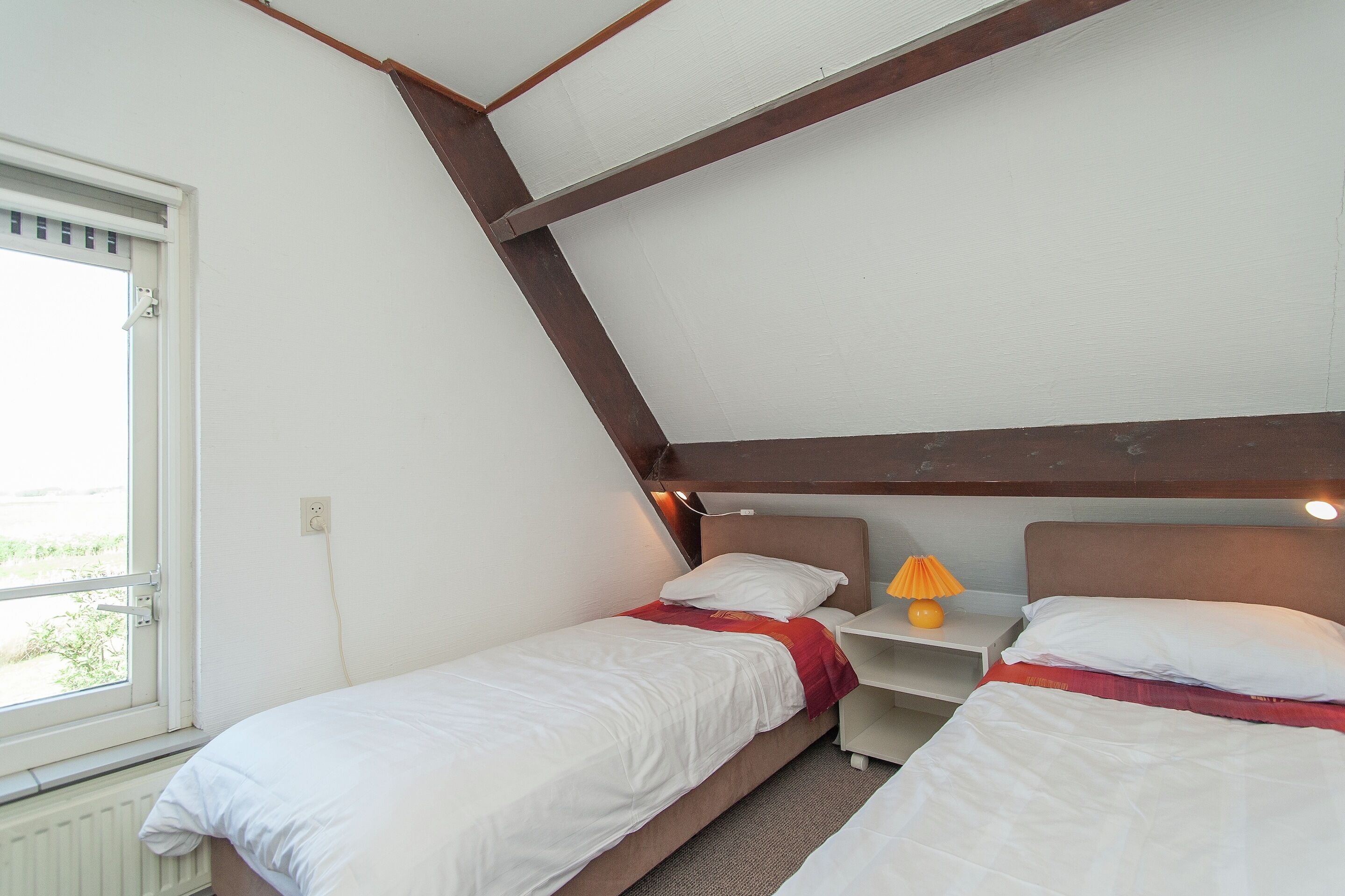 Une chambre avec deux lits simples, une fenêtre, une table de chevet avec une lampe et un plafond en pente.