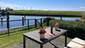 Une table en bois, ornée de plantes en pot et de verres à vin, donnant sur un canal, avec des éoliennes en arrière-plan.