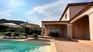 Un área de piscina con sillones de descanso y sombrillas, un patio con mesa y sillas, y un edificio con techo de tejas.