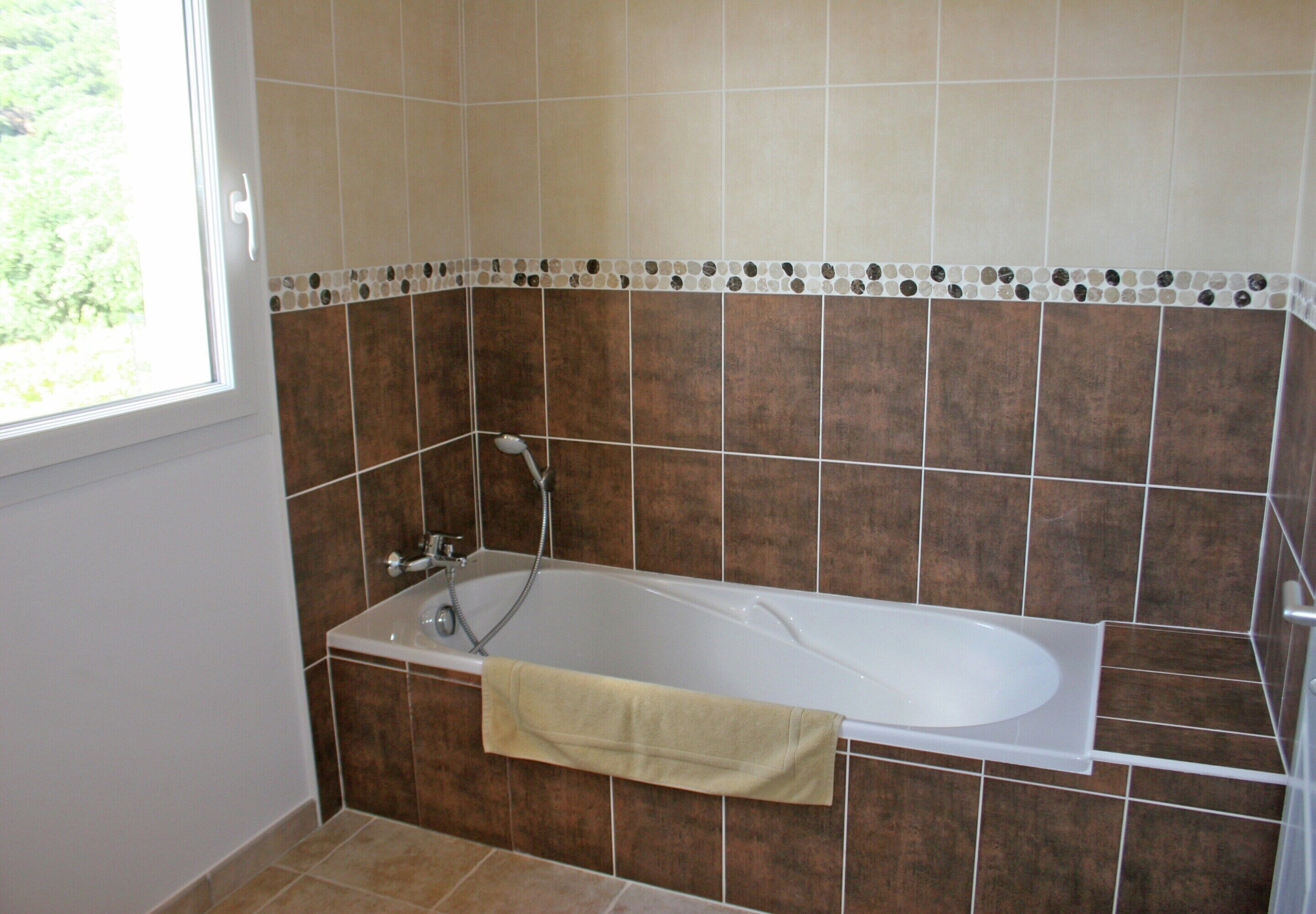 Un baño con bañera, ventana y paredes de baldosas marrones.