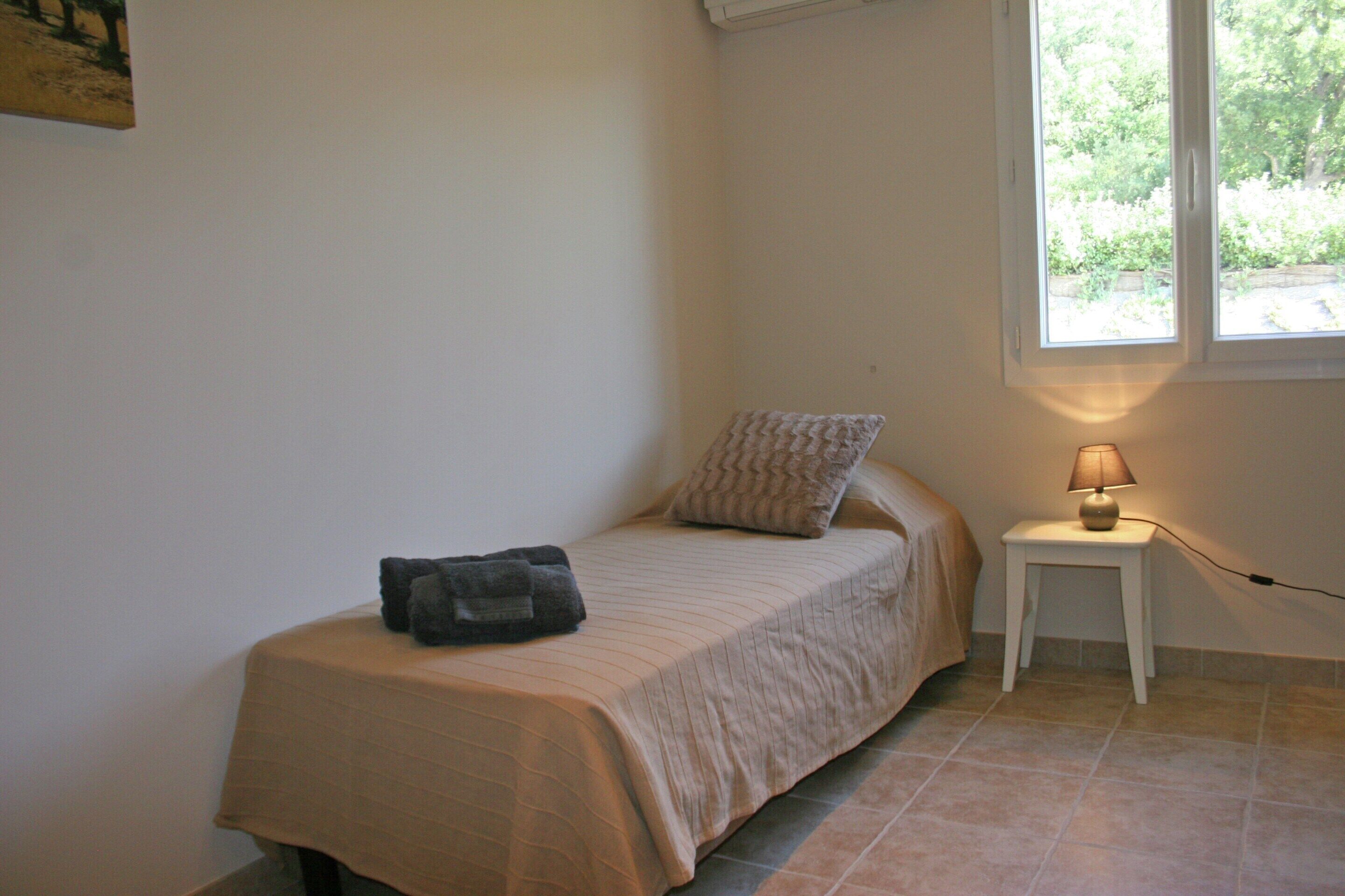 A single bed with a beige bedspread, a pillow, a small bedside table with a lamp, and a window with a view of greenery.