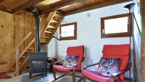 A wooden interior with a wood-burning stove, two red chairs with deer pillows, a wooden staircase, and two windows. - Quiet Chalet With Garden (Saint-Quirin)