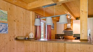 Interior de madera con cocina, dos taburetes de bar y una ventana con cortinas.