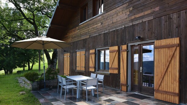 A rustic building with a patio area featuring a table and chairs, a wooden door, and a large umbrella. - Chalet in Saulxures Near Ski Resorts (Saulxures-sur-Moselotte)