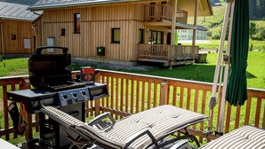 En träaltan med en grill, solstolar och utsikt över ett hus i bakgrunden.