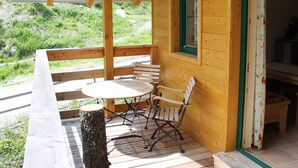Una terraza de madera con una mesa y sillas, una ventana con detalles verdes, y vistas al verdor exterior.