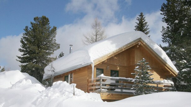 Una cabaña de madera con techo inclinado, rodeada de nieve y árboles.