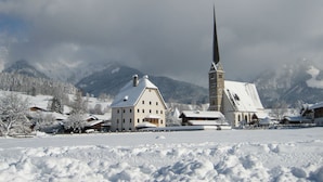 Un village enneigé, avec une église imposante dotée d’un flèche haute, entourée de bâtiments résidentiels et bordé de montagnes en arrière-plan.