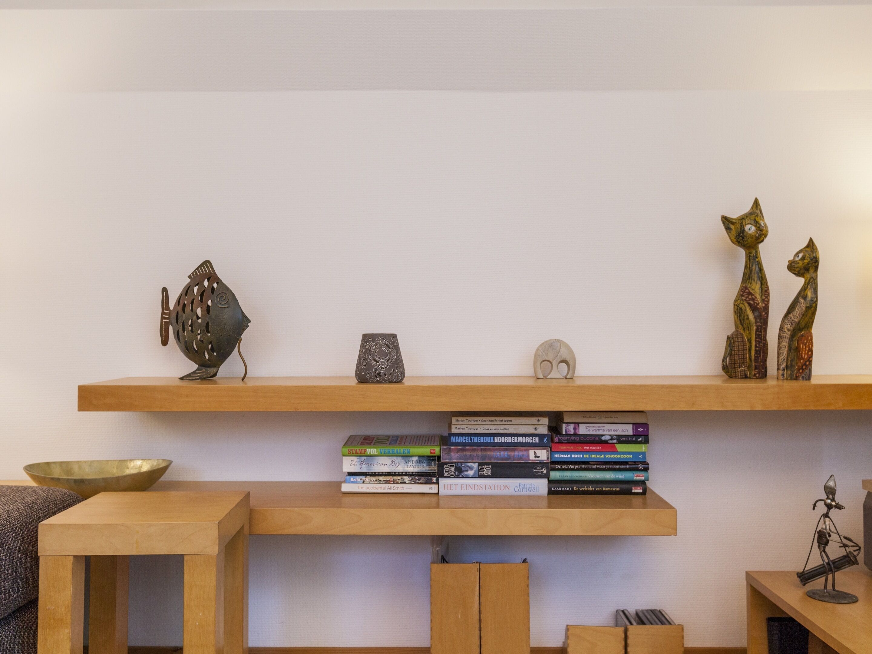 A room with a wooden shelf holding books, a decorative fish, and two cat sculptures.