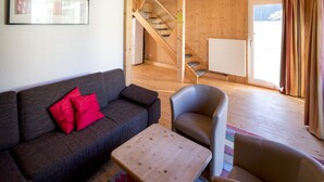 A living room with a sofa, a chair, a small table, a staircase, and a window with curtains. - Classic Chalet With Infrared Sauna (Murau)