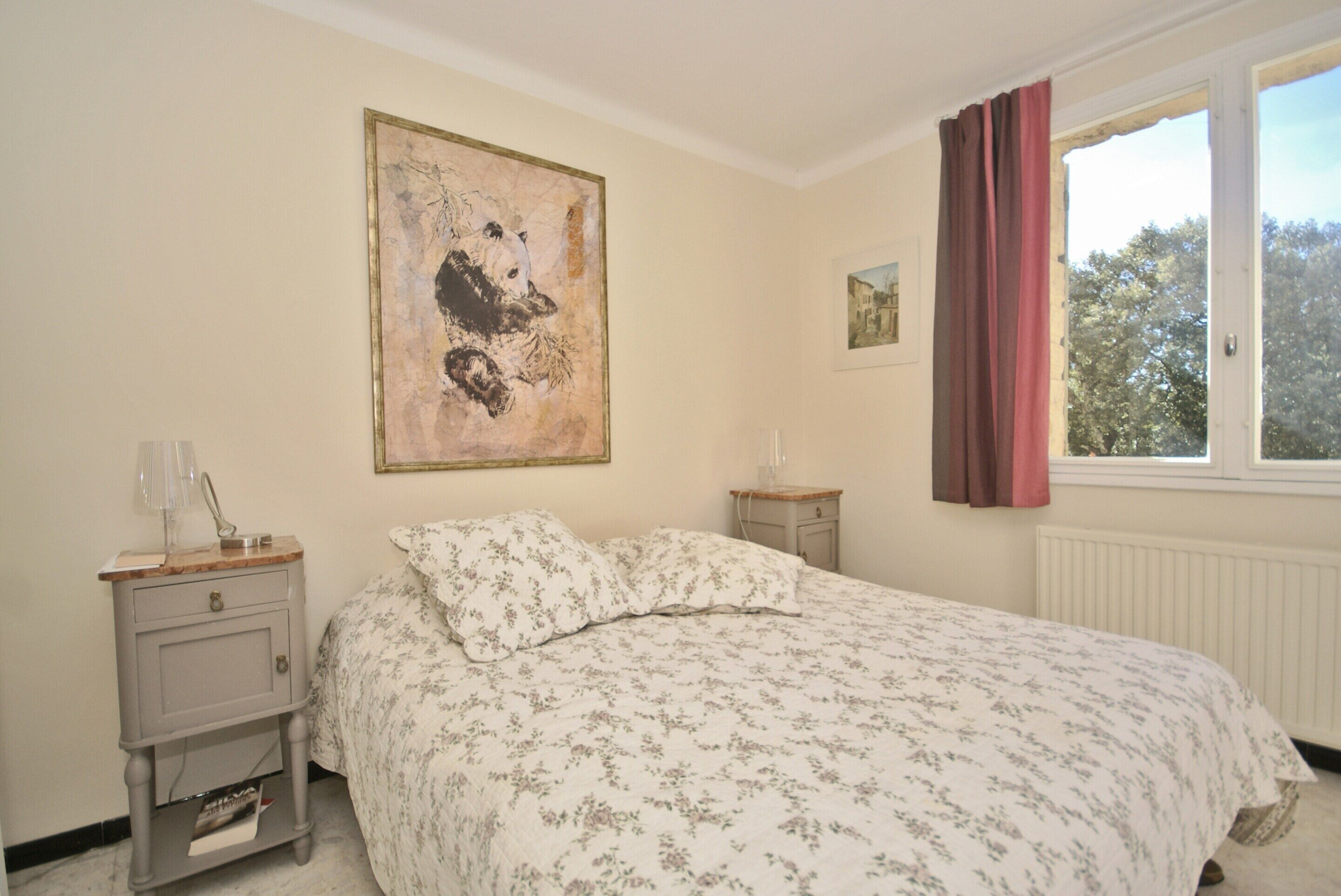 Une chambre à coucher avec un lit, une table de chevet, une lampe, une fenêtre avec des rideaux et un tableau au mur.