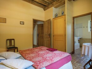 Een slaapkamer met een bed, een stoel, een houten deur en een zichtbare badkamer door een geopende deur.