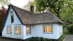 A traditional thatched-roof cottage with white walls and blue trim, featuring three windows with white frames and a small chimney.