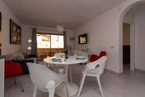 A dining area with a round table, white chairs, and a red chair. There is a sofa and a small table with a lamp in the background. - Mare Verde 95 - One Bed (Costa Adeje)