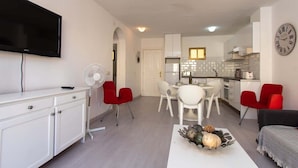 Ein modernes Wohnzimmer mit Flachbildfernseher, einem Essbereich mit weißen Stühlen, einer Küche mit weißen Schränken und einer Uhr an der Wand.