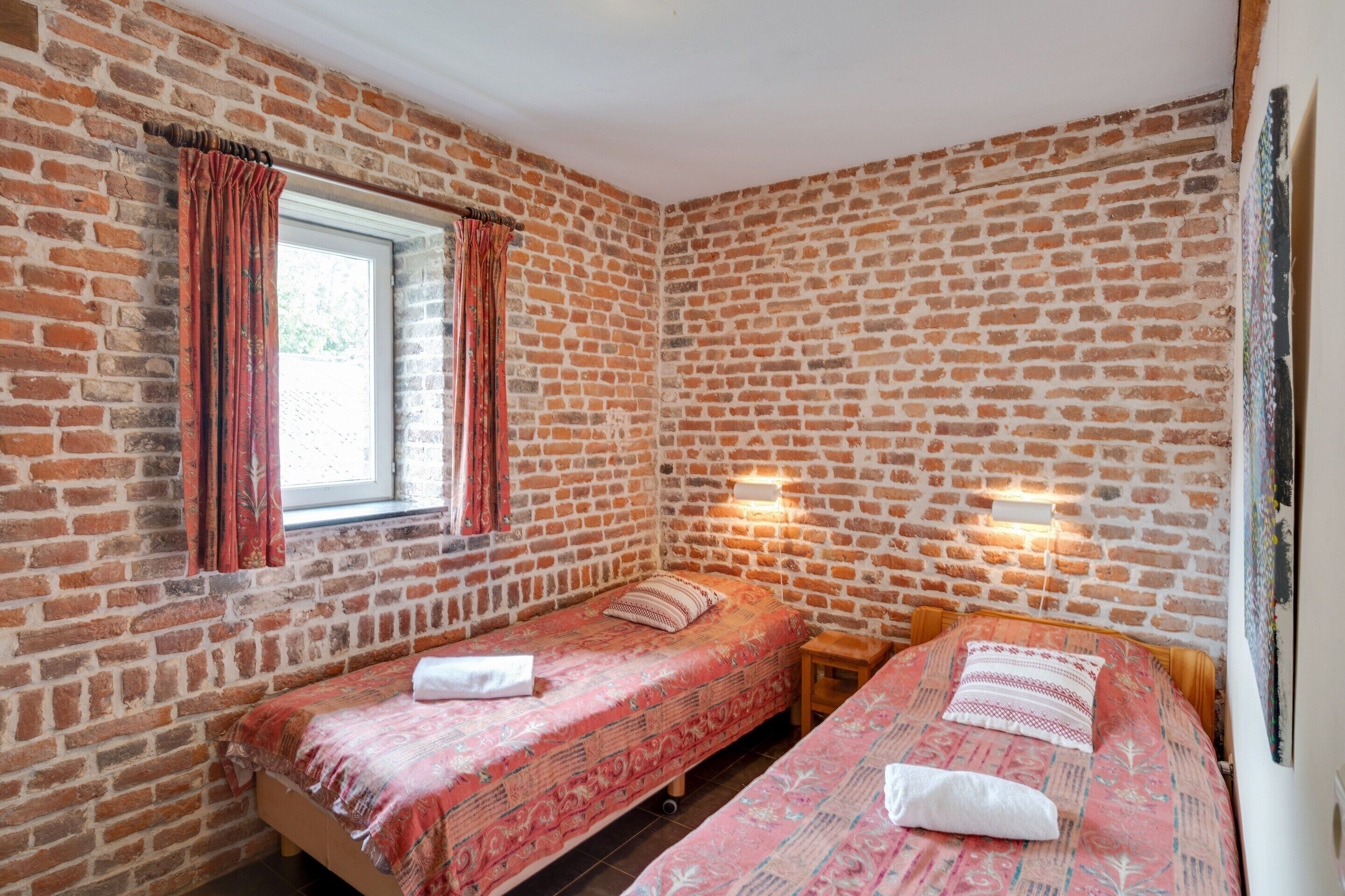 Une chambre avec deux lits, une fenêtre avec des rideaux, des murs en briques et une petite table.