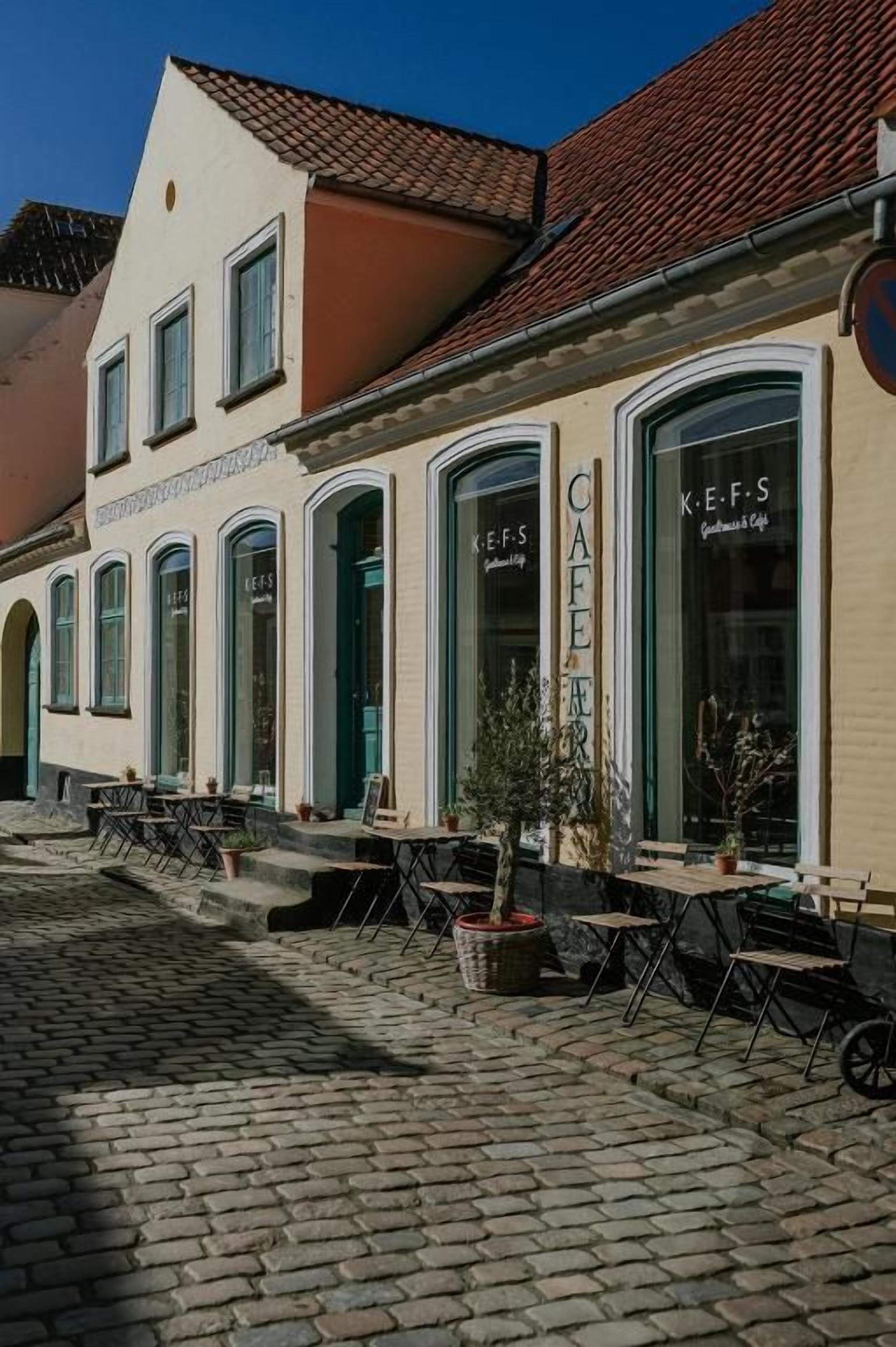 A café with outdoor seating, a cobblestone street, and a building with arched windows.