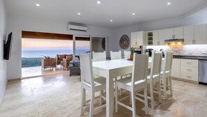 Eine moderne Küche mit weißen Schränken, ein Essbereich mit einem weißen Tisch und Stühlen sowie Blick auf den Ozean.