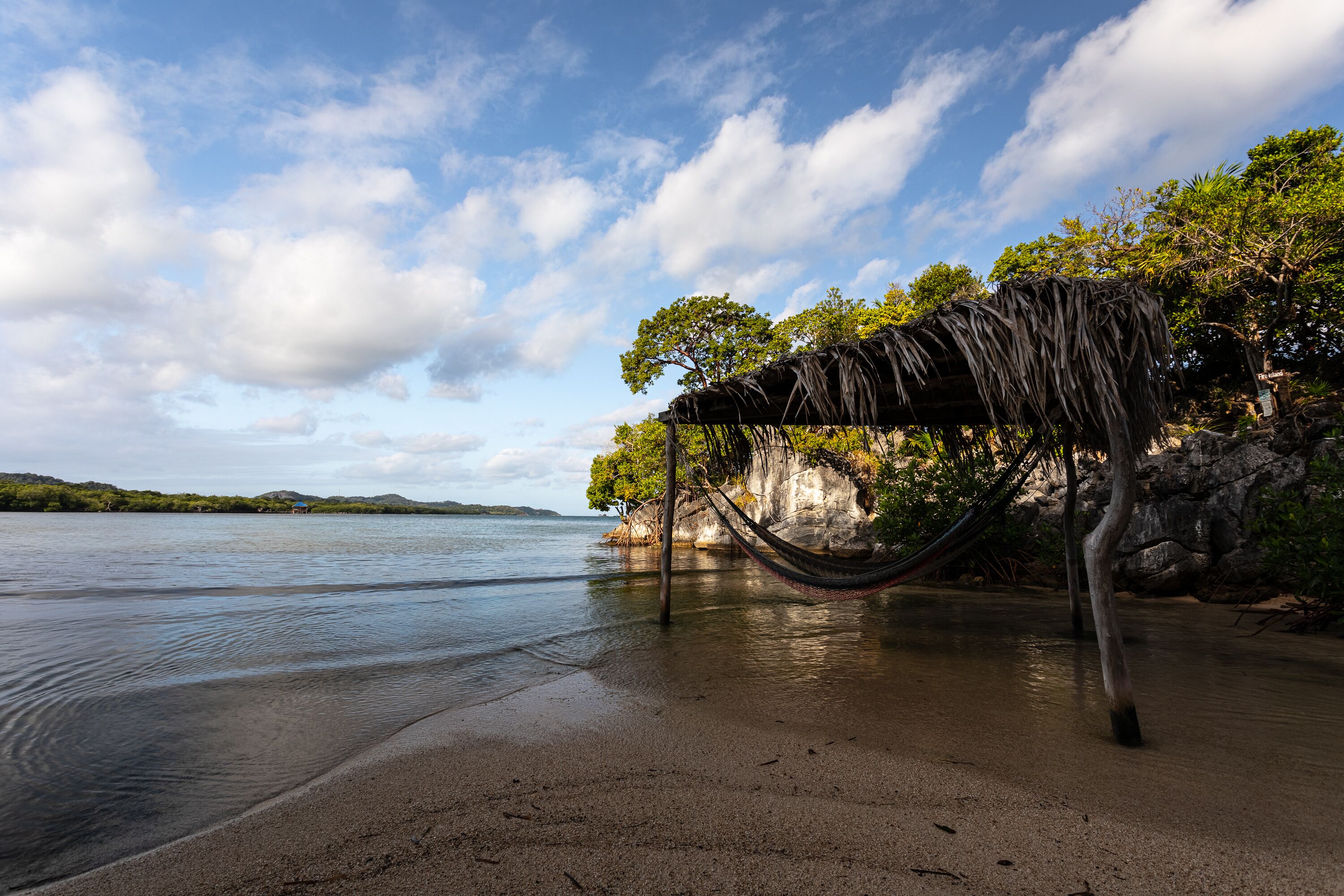 Una hamaca sobre una playa arenosa, con un acantilado rocoso y árboles al fondo.