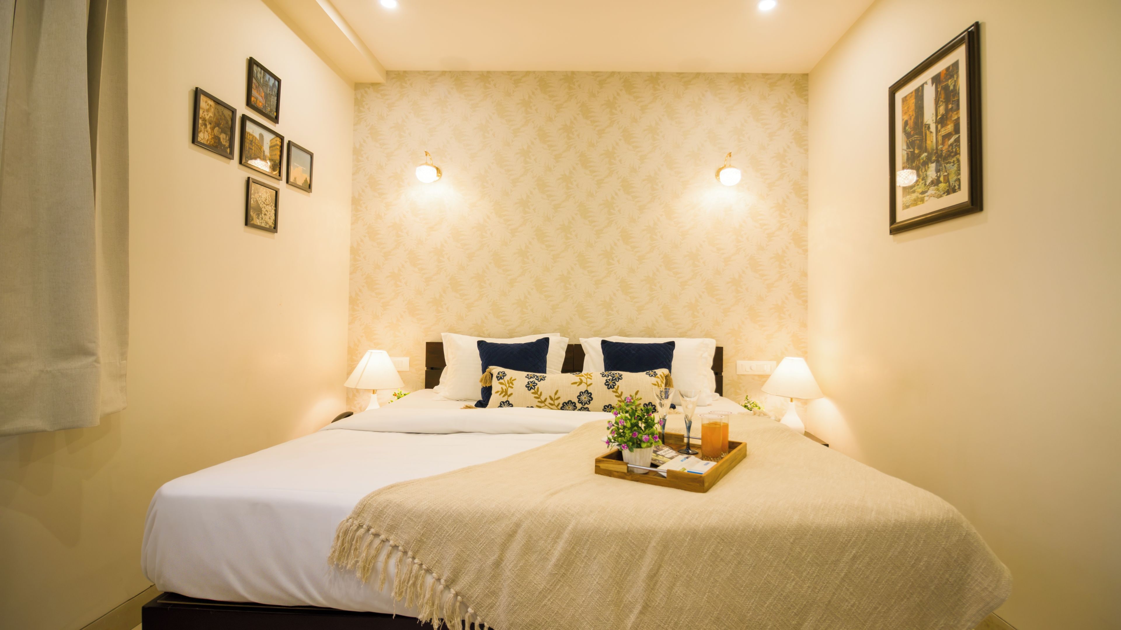 Quarto de hotel com uma cama grande, duas luminárias de cabeceira, um suporte com uma planta e papel de parede estampado.