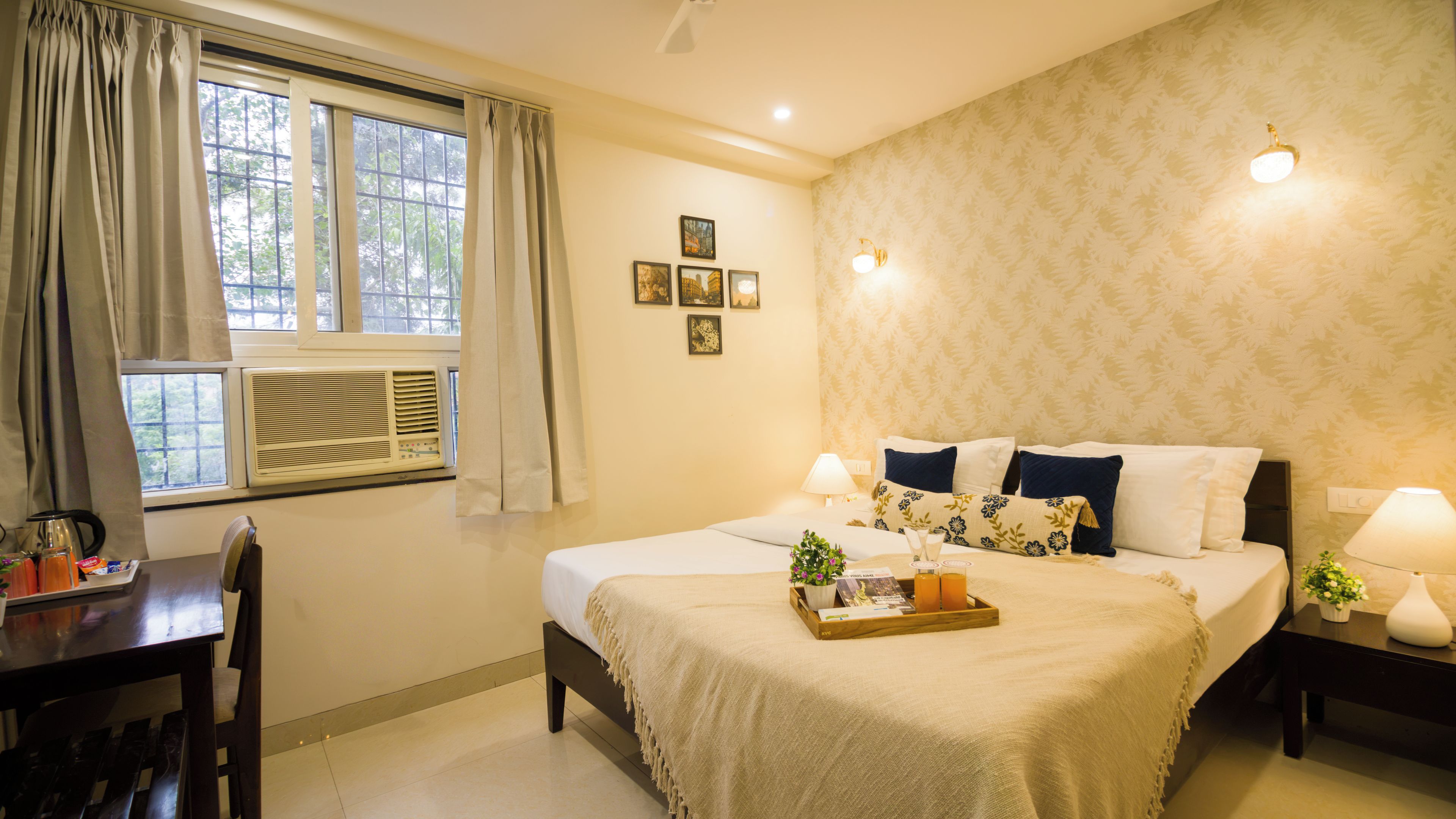 A hotel room with a bed, bedside tables, a desk, an air conditioning unit, and a window with curtains.