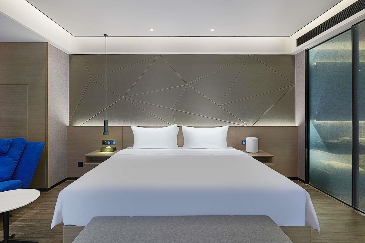 A modern hotel room with a large bed, a bedside table with a lamp, a digital clock, and a bedside heater.
