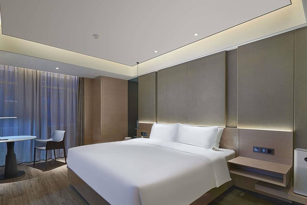 A modern hotel room with a large bed, a bedside table, a chair, and a small table with a lamp.