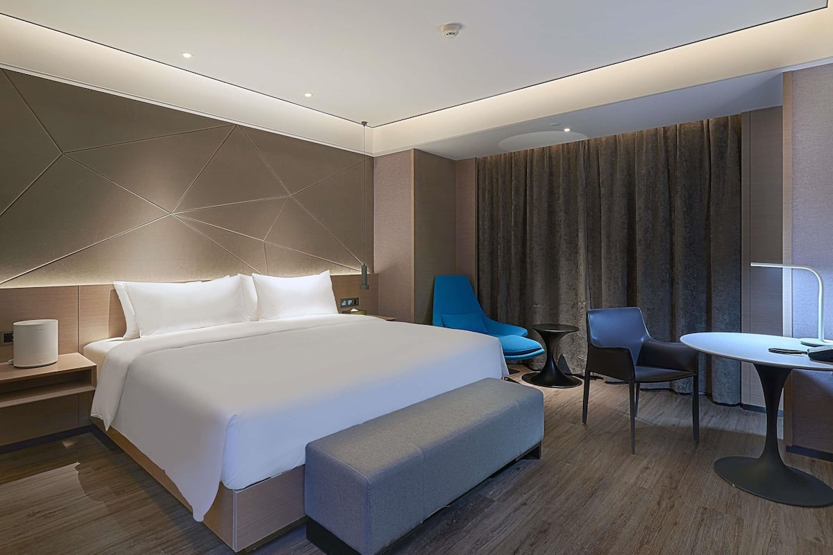 A modern hotel room with a large bed, a desk, a chair, and a small table.