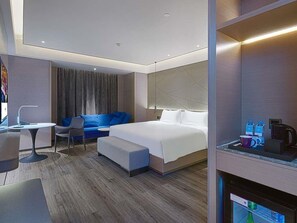 Et moderne hotelværelse med seng, siddeplads med blå sofa, lille bord og kaffemaskine.