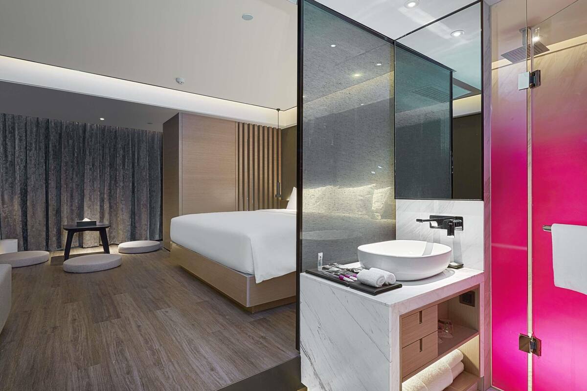 A modern hotel room with a bed, bedside table, and a bathroom with a large mirror, sink, and shower.