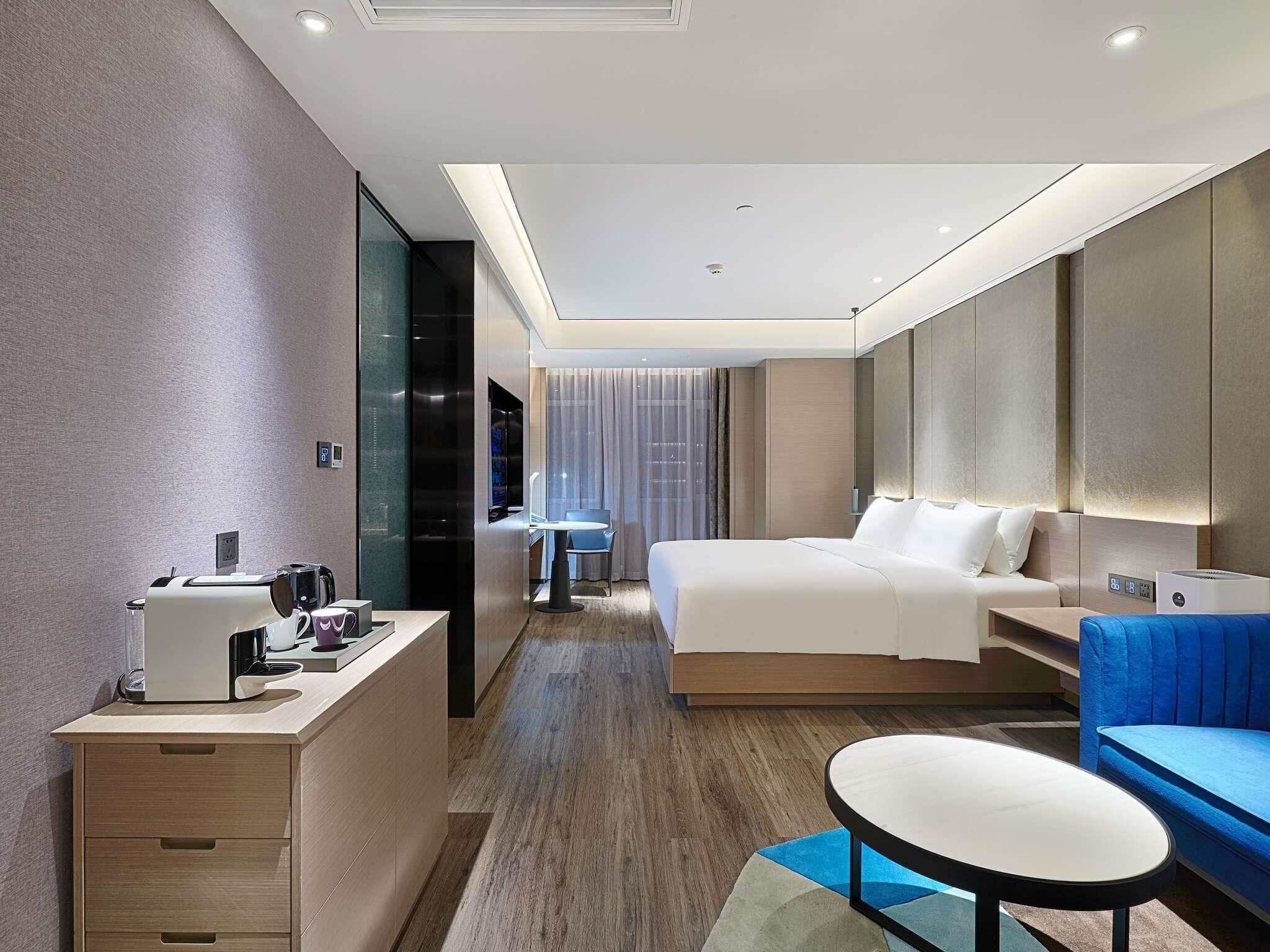 Ein modernes Hotelzimmer mit Bett, Schreibtisch mit Kaffeemaschine, einer Sitzecke mit blauem Sofa und einem großen Spiegel.