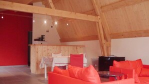 A spacious loft with a high ceiling, a red wall, and a wooden ceiling. There is a dining table with chairs and a sofa with orange cushions. - Holiday Home in Ardeche Near Chateau de Ventadour (Lalevade-d'Ardeche)