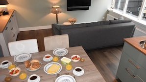A modern living room with a grey sofa, a dining table set with breakfast, a TV mounted on the wall, and a window with a view. - Holiday Home in Egmond by the Beach (Egmond aan Zee)