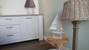 A room with a wooden cabinet, a small round table with a model sailboat, a wicker lamp, and a white textured stool. - Holiday Home in Egmond by the Beach (Egmond aan Zee)