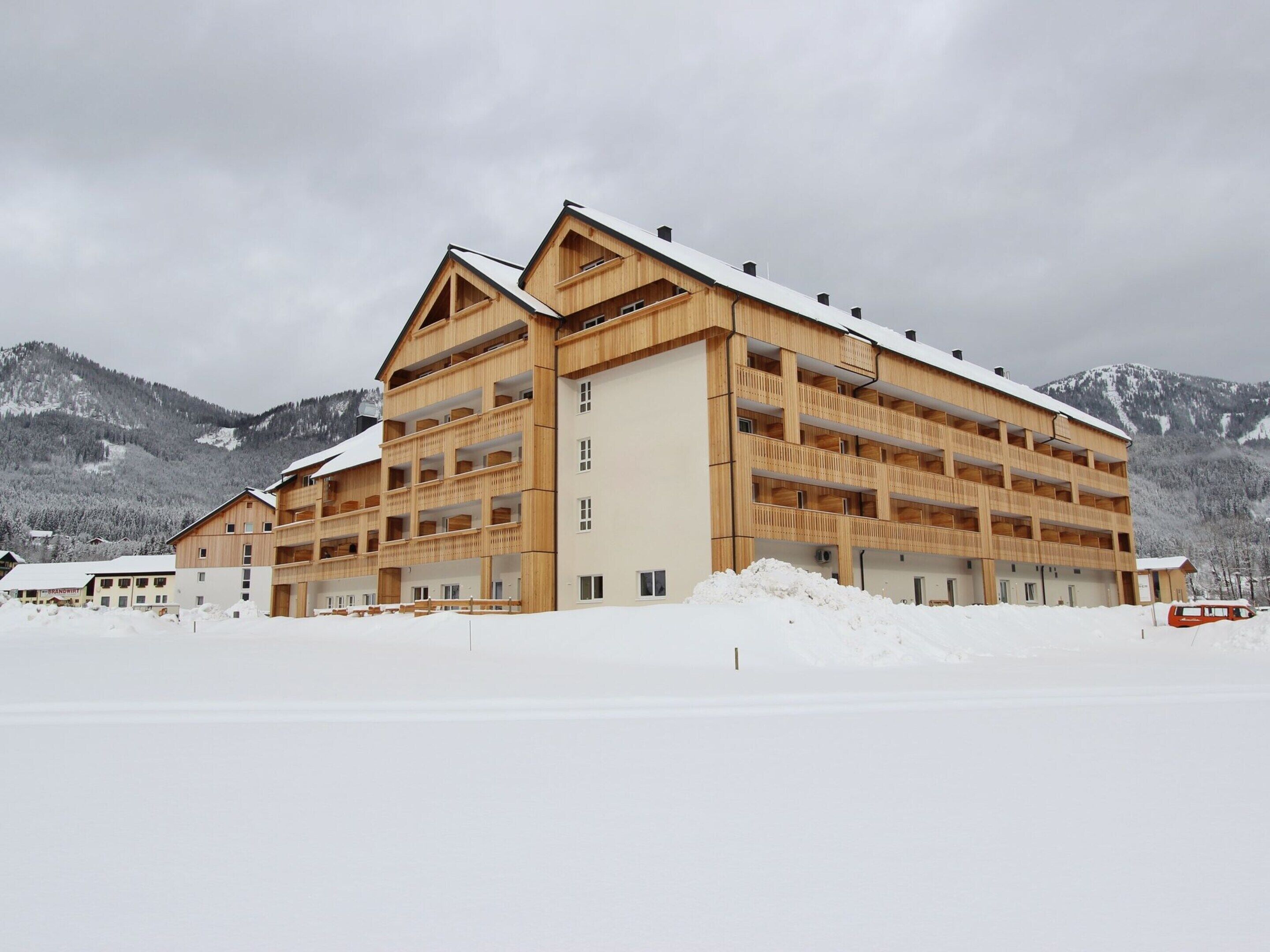 A multi-story wooden building with balconies, set against a snowy mountainous backdrop.
