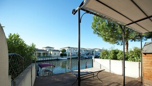 Um deck de madeira com guarda-corpo de metal, uma área coberta com pérgola e vista para uma marina com barcos e edifícios ao fundo.