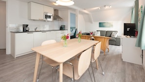 A modern kitchen with white cabinets, a microwave, and a coffee maker. There is a dining area with a wooden table and chairs, and a living area with a sofa and a TV. - Quaint Apartment in Schoorl near Tennis Court (Schoorl)
