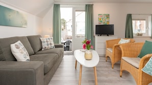 A living room with a grey sofa, a white coffee table, wicker chairs, and a TV on a stand. - Quaint Apartment in Schoorl near Tennis Court (Schoorl)