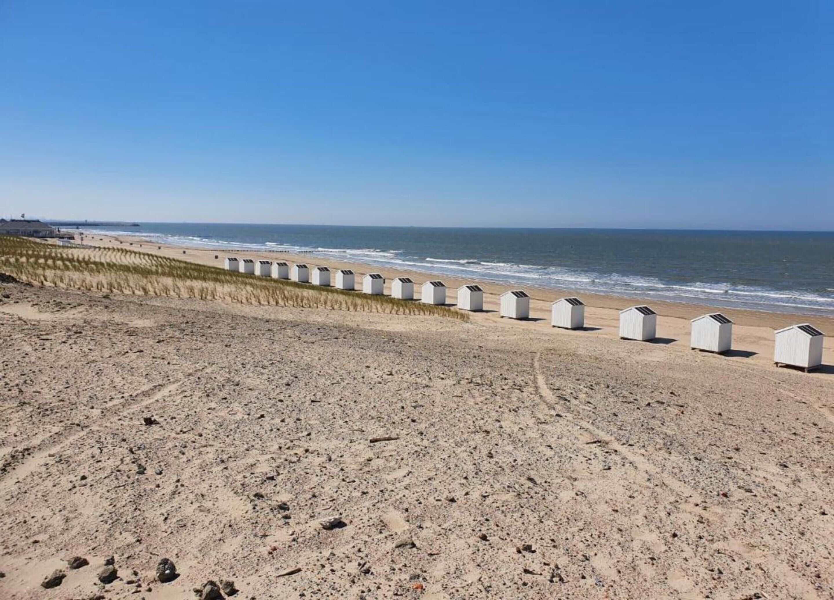 Une plage de sable bordée d’une rangée de cabanes de plage blanches, sous un ciel bleu et dégagé.