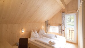 Een slaapkamer in een houten hutje, met een bed, kussens, een bankje en een raam met gordijnen.