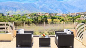 Une terrasse meublée de mobilier en osier, une table avec des fruits et une tasse, et une vue sur une ville située dans un paysage montagneux.