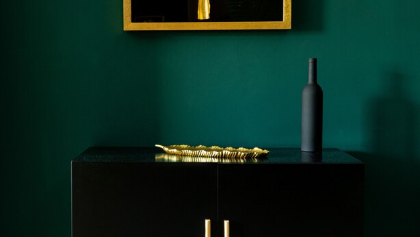 A black cabinet with wooden legs, a gold-framed mirror, and a black vase on top. - Welcome to the Jungle (Bucharest)