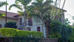 Une maison à deux étages dotée d’un balcon, entourée d’une végétation luxuriante et d’un mur de pierre.
