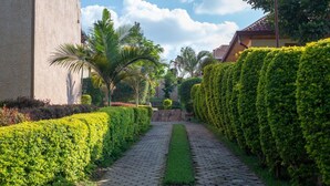 Un chemin pavé, bordé de haies soigneusement taillées et de palmiers, mène à un bâtiment dont le toit est recouvert de tuiles.