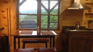 Interior de uma cabana de madeira com mesa de jantar e bancos, cozinha com fogão e chaleira, e janela com persianas dando para o verde.